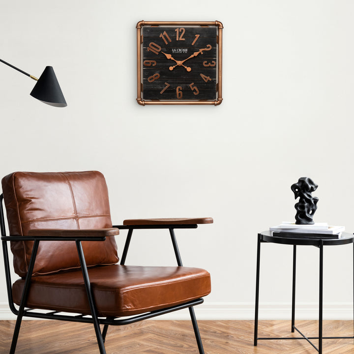 Lifestyle image of the 404-3412D 16-inch Foundry Metal &amp; Wood Wall Clock - Dark, shown hanging on a white wall above a cognac leather chair and side table. The clock features a square metal pipe frame in a copper finish, a black-stained wood slat clock face with copper-finished metal numbers and hands.