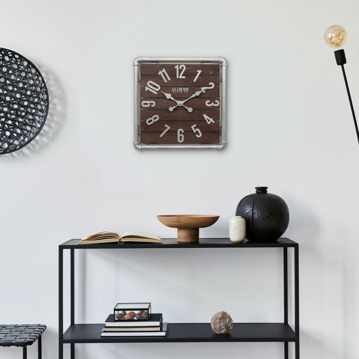 Lifestyle image of the 404-3412L 16-inch Foundry Metal &amp; Wood Wall Clock - Light, shown hanging above a black metal shelving unit with books and vases. The clock features a square metal pipe frame in a weathered pewter finish, a truffle-brown-stained wood slat clock face with pewter-finished metal numbers and hands.