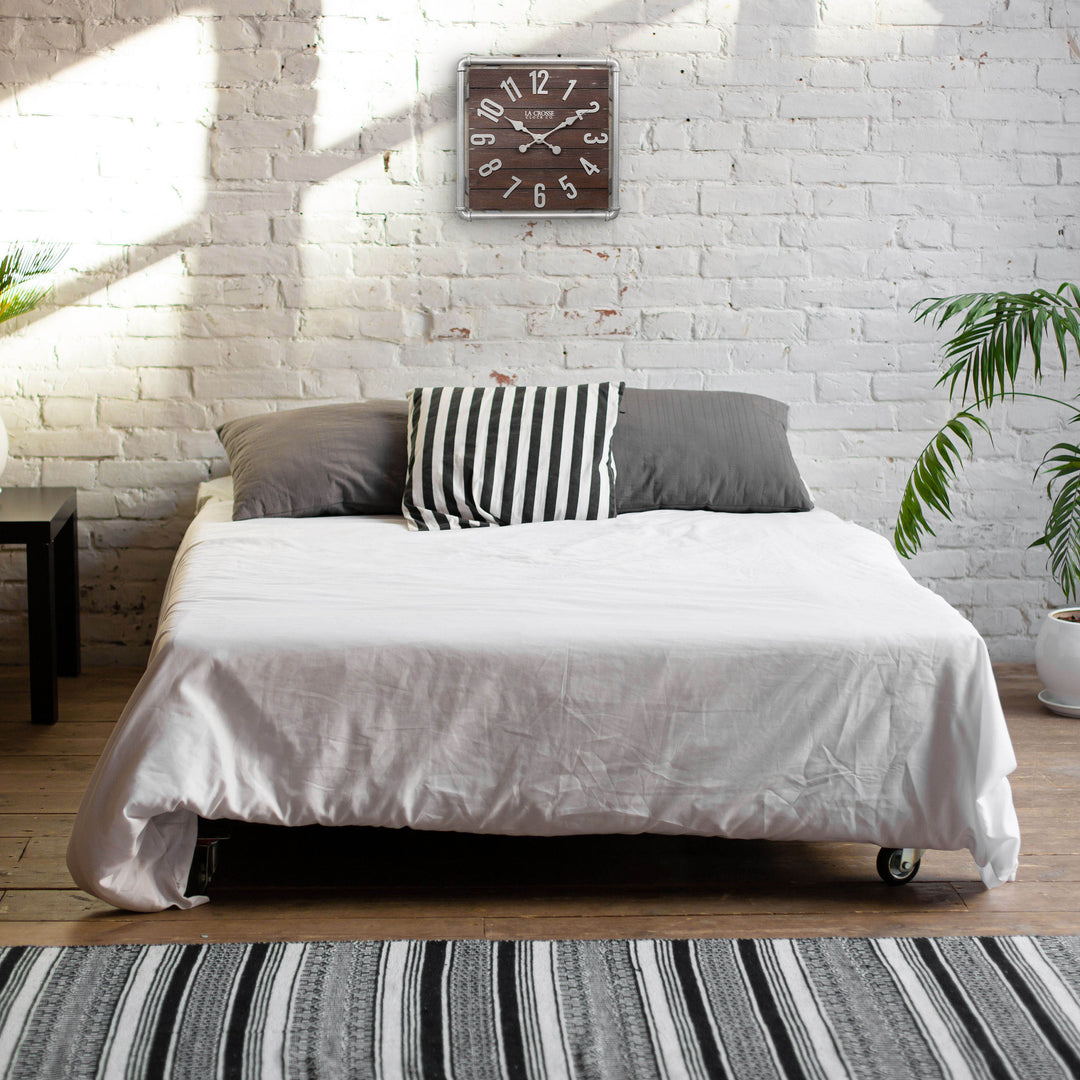 Lifestyle image of the 404-3412L 16-inch Foundry Metal &amp; Wood Wall Clock - Light, shown hanging on a white-washed brick wall above a bed. The clock features a square metal pipe frame in a weathered pewter finish, a truffle-brown-stained wood slat clock face with pewter-finished metal numbers and hands.
