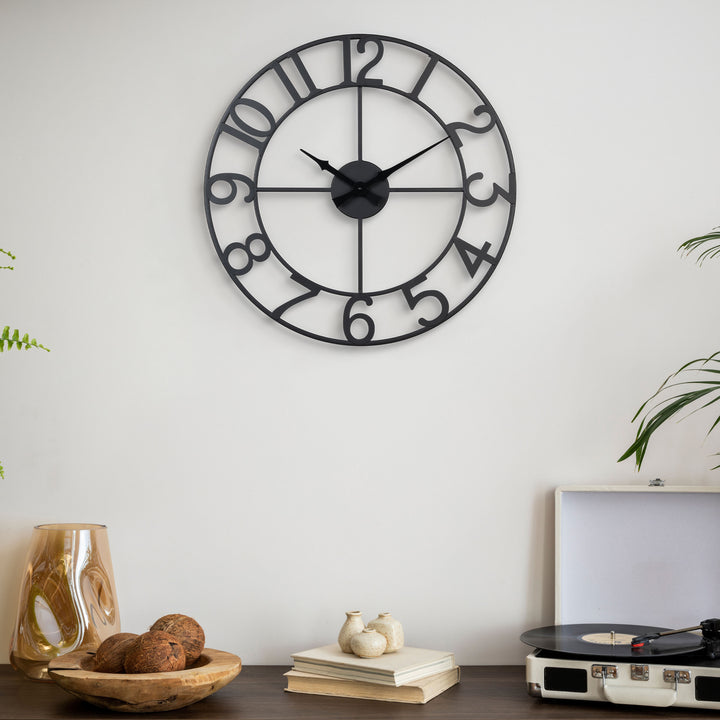 Lifestyle image of the 404-3451A 20-inch Metal Tower Wall Clock - Arabic hanging on a light coloroed wall above a side table with a vintage record player, sculptures, books, and a vase. This skeleton tower clock features Arabic numbers, hour and minute hands all constructed in metal.