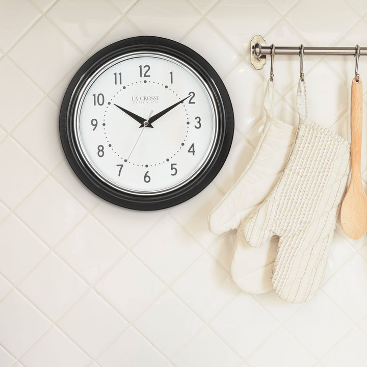 Lifestyle image of the 404-3624B 9.5-inch Diner Wall Clock hanging on a white tile wall in a kitchen. The clock features a black frame with a chrome bezel accent, an off-white dial with black numbers and hour dots, a silver second hand, and black hour and minute hands.
