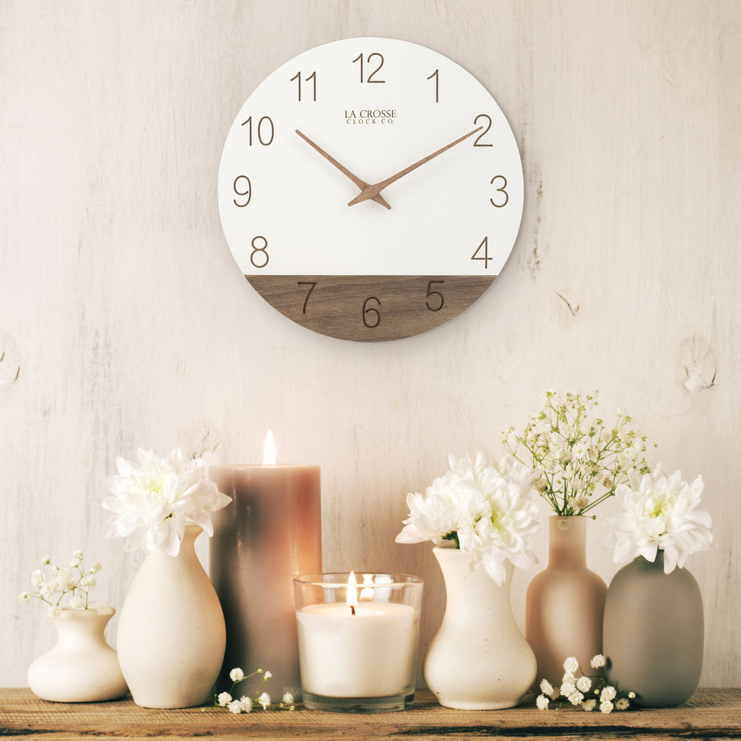 Lifestyle image of the 404-3630B 12-inch Sierra Wood Wall - White, hanging on a cream paneled wall above a wooden rustic shelf holding vases of flowers and candles. The clock features laser-cut numbers, a black and wood open-face dial, and wood hour and minute hands.