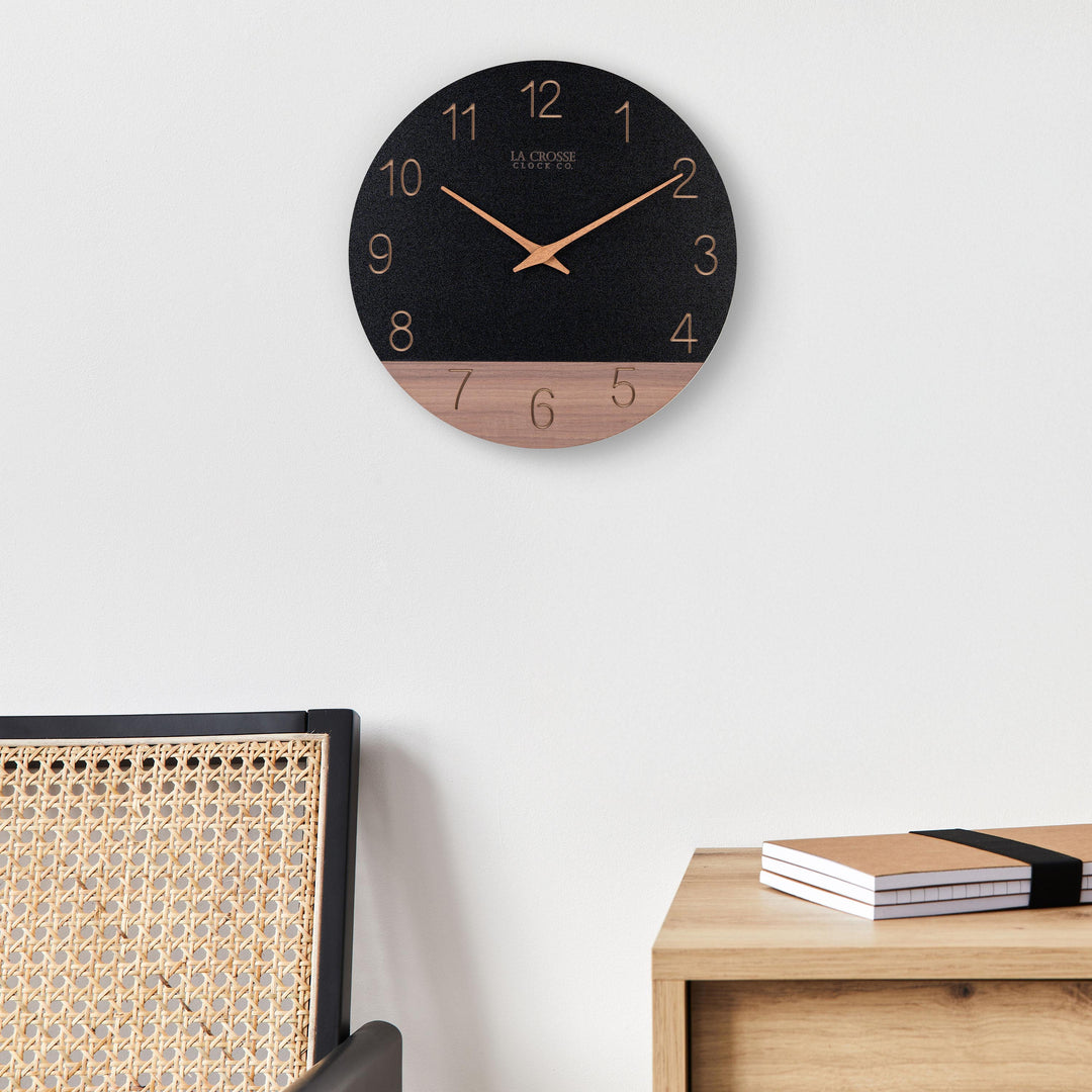 Lifestyle image of the 404-3630C 12-inch Sierra Wood Wall Clock - Black, the clock is shown hanging on a wall above a side table and a wicker chair. The clock features a black and wood split design open-face dial, with laser cut numbers, and wood hour and minute hands.