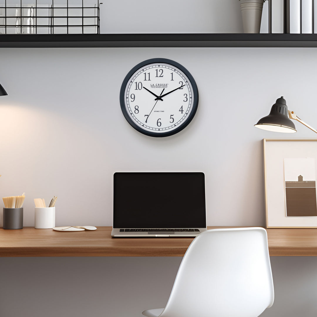 Lifestyle image of the 404-3826 10-inch Atomic Analog Wall Clock hanging on a wall above a desk with a laptop, desk accessories, and a chair. The clock features a black flush-mount frame, stylized black numerals, and black metal hands
