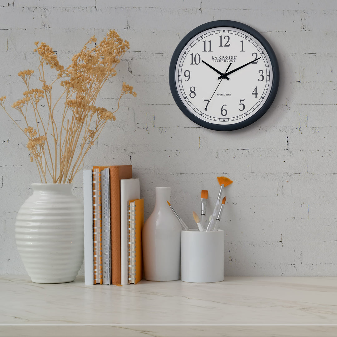 Lifestyle image of the 404-3826 10-inch Atomic Analog Wall Clock hanging on a white-washed brick wall above a marble countertop with books and white vases. The clock features a black flush-mount frame, stylized black numerals, and black metal hands.