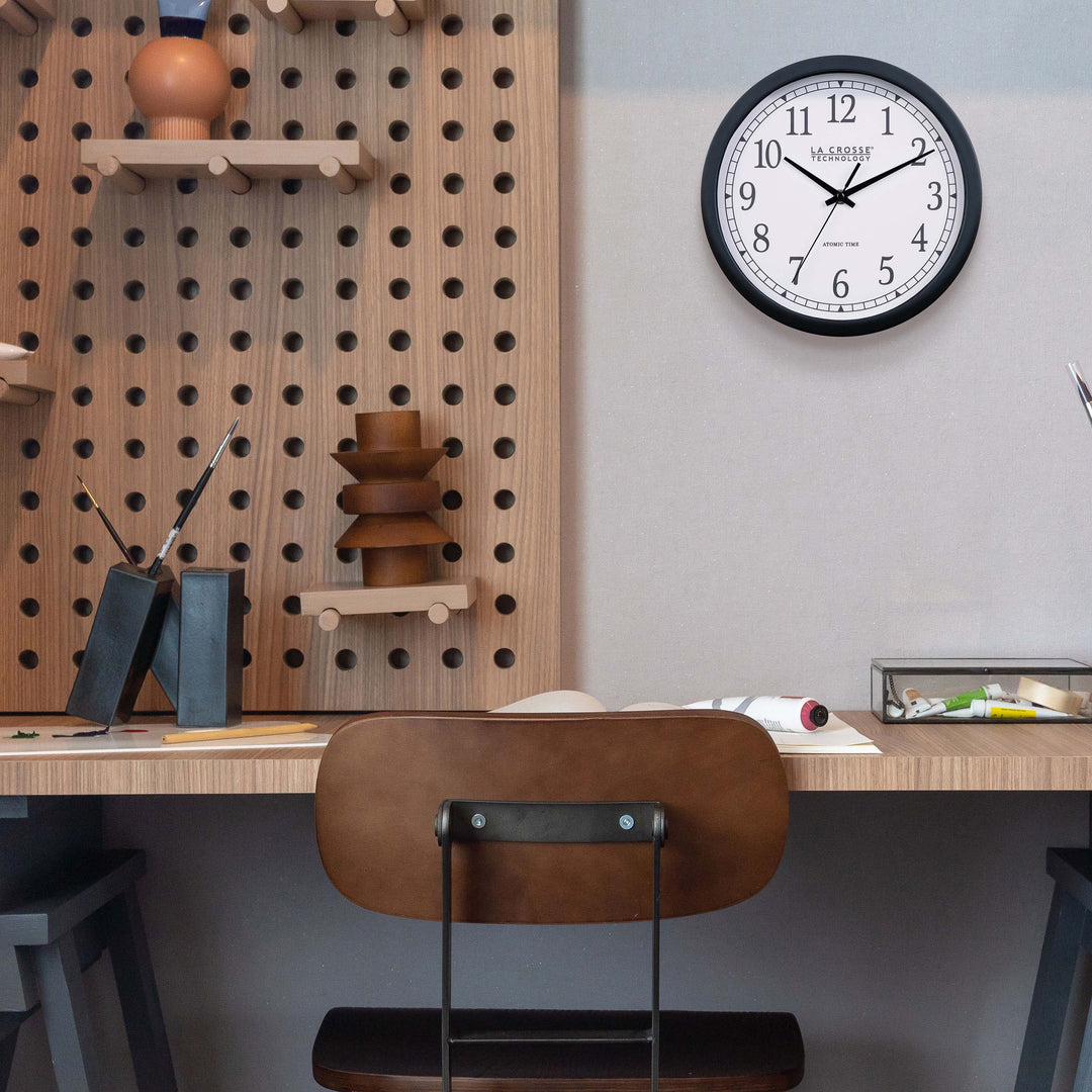 Lifestyle image of the 404-3826 10-inch Atomic Analog Wall Clock hanging on a white wall above a desk with an architectural design. The clock features a black flush-mount frame, stylized black numerals, and black metal hands.