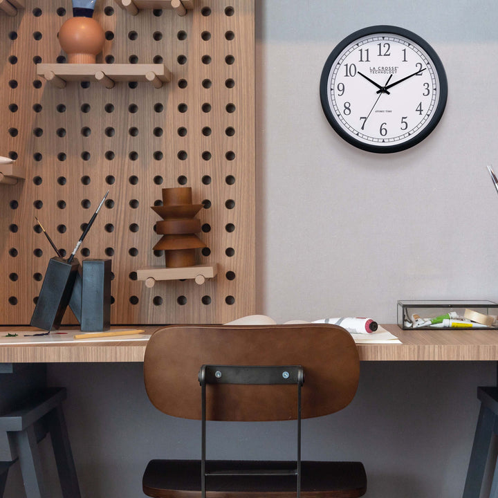 Lifestyle image of the 404-3826 10-inch Atomic Analog Wall Clock hanging on a white wall above a desk with an architectural design. The clock features a black flush-mount frame, stylized black numerals, and black metal hands.