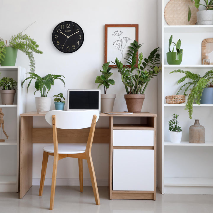 Lifestyle image of the 404-3831B 12-inch Paxton Wall Clock - Black, with Silent Sweep, shown hanging on a wall in a home office with a desk, bookshelves, and plants. The clock features a flush-mount black frame, a matching color dial, brass raised numbers, and brass-finished hour, minute, and second hands.