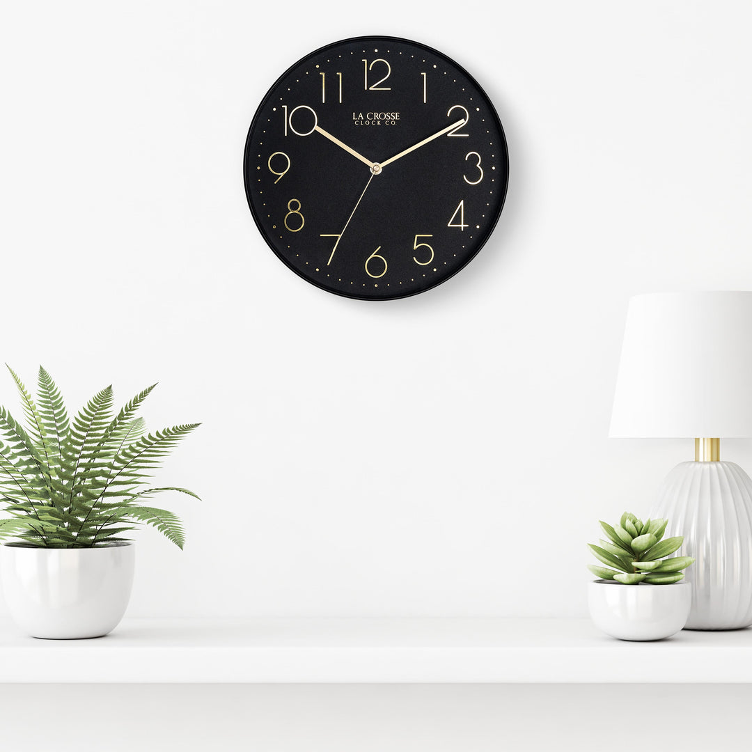 Lifestyle image of the 404-3831B 12-inch Paxton Wall Clock - Black, with Silent Sweep, hanging on a white wall above a white shelf with a white lamp and plants in white vases. The clock features a flush-mount black frame, a matching color dial, brass raised numbers, and brass-finished hour, minute, and second hands.