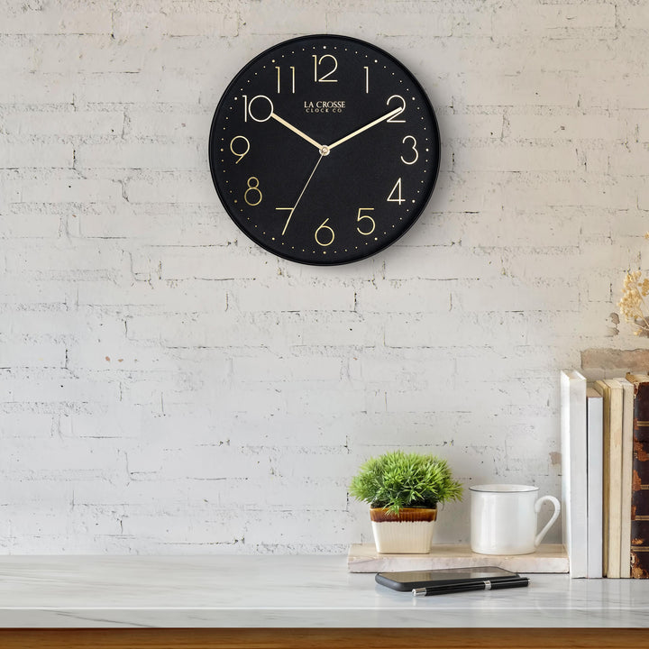 Lifestyle image of the 404-3831B 12-inch Paxton Wall Clock - Black, with Silent Sweep, hanging on a white-washed brick wall above a marble countertop with books, a plant, and a phone. The clock features a flush-mount black frame, a matching color dial, brass raised numbers, and brass-finished hour, minute, and second hands.