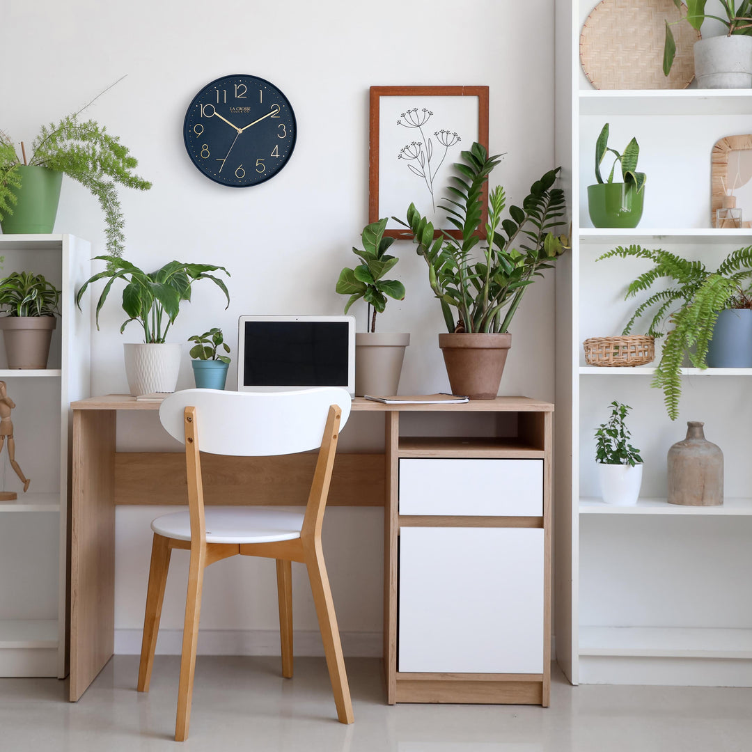Lifestyle image of the 404-3831NB 12-inch Paxton Wall Clock - Navy Blue, with Silent Sweep, shown hanging on a wall in a home office with a desk, bookshelves, and plants. The clock features a flush-mount navy blue frame, a matching color dial, brass raised numbers, and brass-finished hour, minute, and second hands.