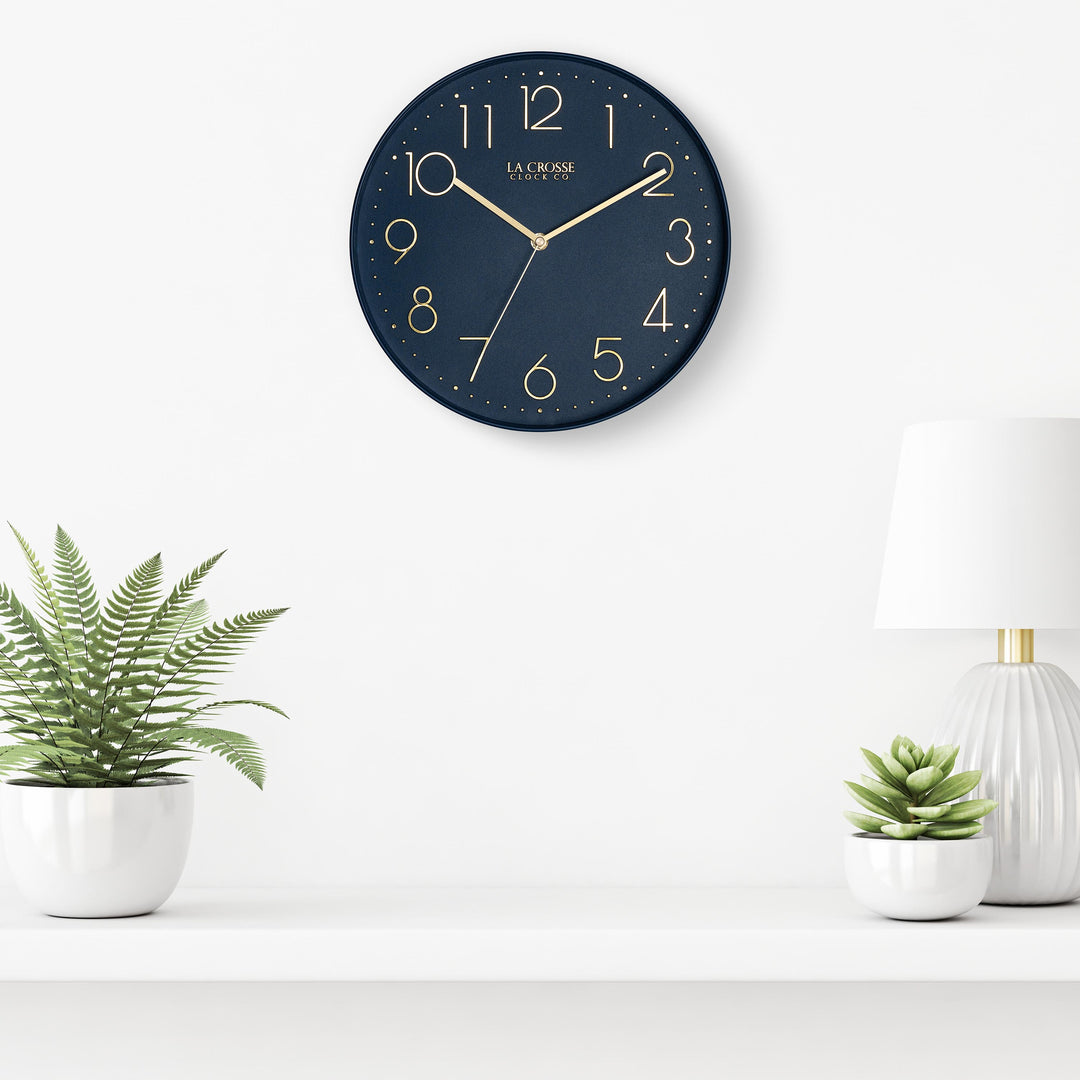 Lifestyle image of the 404-3831NB 12-inch Paxton Wall Clock - Navy Blue, with Silent Sweep, hanging on a white wall above a white shelf with a white lamp and plants in white vases. The clock features a flush-mount navy blue frame, a matching color dial, brass raised numbers, and brass-finished hour, minute, and second hands.