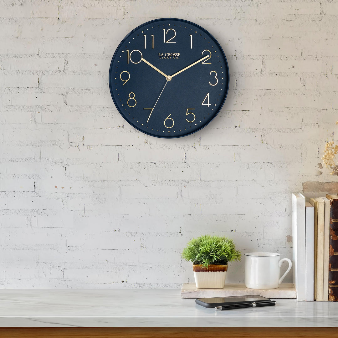 Lifestyle image of the 404-3831NB 12-inch Paxton Wall Clock - Navy Blue, with Silent Sweep, hanging on a white-washed brick wall above a marble countertop with books, a plant, and a phone. The clock features a flush-mount navy blue frame, a matching color dial, brass raised numbers, and brass-finished hour, minute, and second hands.
