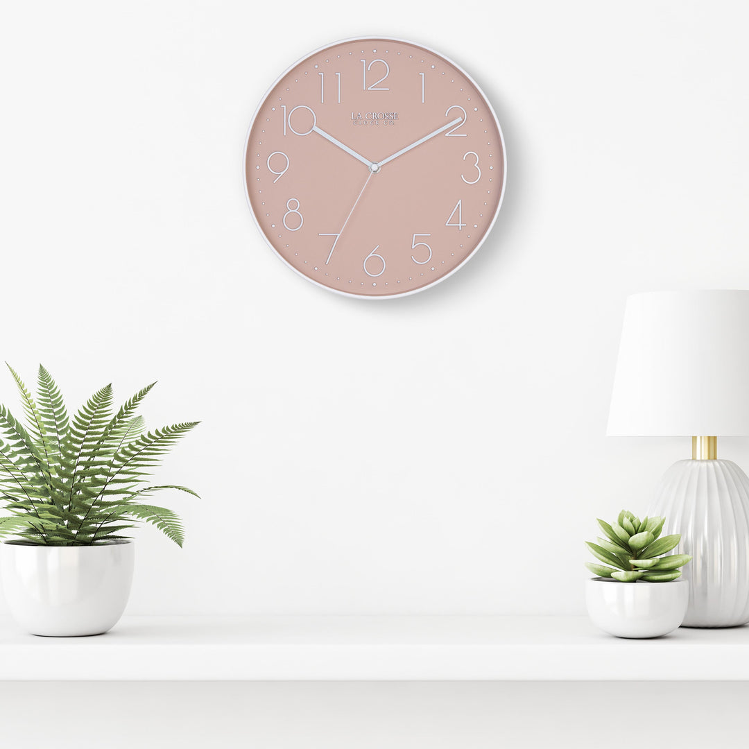 Lifestyle image of the 404-3831P 12-inch Paxton Wall Clock - Peach, with Silent Sweep, hanging on a white wall above a white shelf with a white lamp and plants in white vases. The clock features a flush-mount peach frame, a matching color dial, white raised numbers, and white metal hour, minute, and second hands.