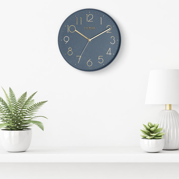 Lifestyle image of the 404-3831SP 12-inch Paxton Wall Clock - Speckled, with Silent Sweep, hanging on a white wall above a white shelf with a white lamp and plants in white vases. The clock features a flush-mount speckled blue frame, a matching color dial, brass raised numbers, and brass-finished hour, minute, and second hands.
