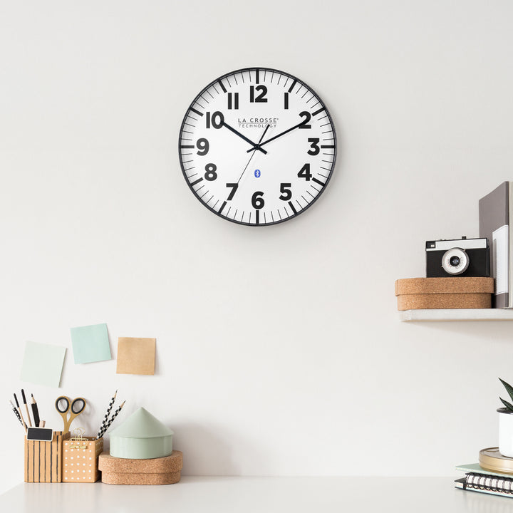 Lifestyle image of the 404-4536 14.2-inch Insta-Set Wall Clock with Bluetooth® Technology shown hanging on a white wall above a white desk with desk accessories and a shelf holding books, a cork box, and a camera. The clock features a white dial with bold black numbers and markings, a black flush-mount frame, and black hands.