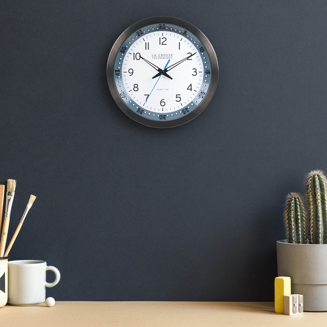 Lifestyle image of the 404-54667 10-inch Atomic Metal Wall Clock with Blue Chapter Ring hanging on a navy wall over a light wood color desk with a cup, paint brushes, and a cactus. The clock features a black stainless steel frame with a blue chapter ring and raised seconds indicators, surrounding a white dial with black numbers, metal hands, and a blue accent second hand.