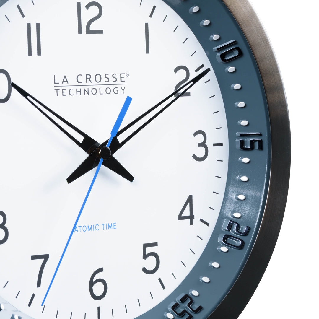 Close-up image of the dial of the 404-54667 10-inch Atomic Metal Wall Clock with Blue Chapter Ring. The clock features a black stainless steel frame with a blue chapter ring and raised seconds indicators, surrounding a white dial with black numbers, metal hands, and a blue accent second hand.