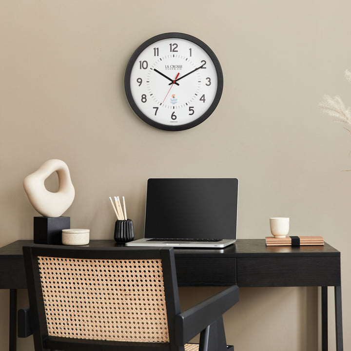 Lifestyle image of the 404-5537 14.5-inch Wall Clock hanging on a wall over a black desk, rattan chair, with a lap top, sculpture and desk accessories. The clock features a black flush mount frame, black bold numbers, black hour and minute hands, and a red second hand with two logos on the clock face, La Crosse Clock Co. and Chicago Lighthouse industries.