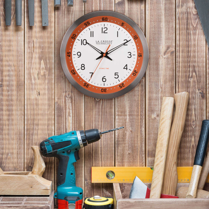 Lifestyle image of the 404-87696 10-inch Atomic Metal Wall Clock - Chapter Ring: Orange shown hanging at a workbench with tools. The clock features a white dial, an orange chapter ring with seconds indicator, black hands, an orange second hand, and a stainless steel flush-mount frame.