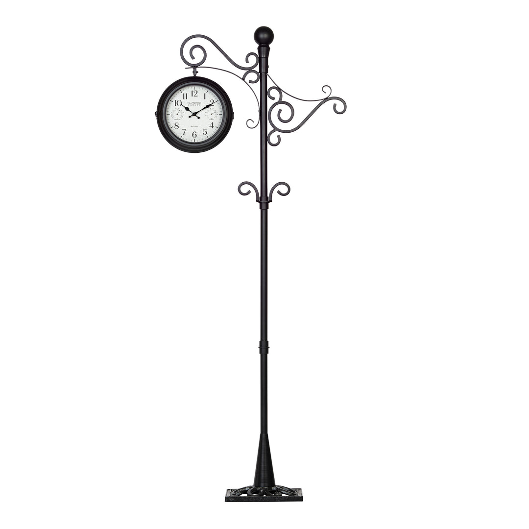 Full head-on image of the 425-34188 Outdoor Dual-Sided Metal Pedestal Clock. The clock features a temperature dial in a Fahrenheit scale, a humidity dial, decorative black metal hands, black numbers on a flecked white dial surrounded by a dark brown metal frame., with a 72-inch decorative scrollwork pedestal with a plant hanger.