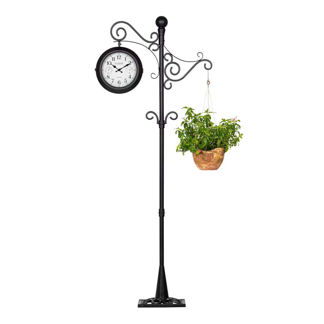 Full head-on image of the 425-34188 Outdoor Dual-Sided Metal Pedestal Clock shown with a hanging flower basket off the plant hook. The clock features a temperature dial in a Fahrenheit scale, a humidity dial, decorative black metal hands, black numbers on a flecked white dial surrounded by a dark brown metal frame., with a 72-inch decorative scrollwork pedestal with a plant hanger.