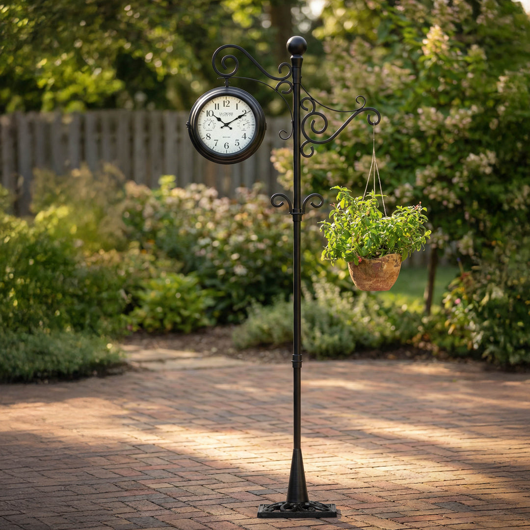 Lifestyle image of the 425-34188 Outdoor Dual-Sided Metal Pedestal Clock shown utilizing the hanging basket hook on a brick patio with the yard and fence in the background. The clock features a temperature dial in a Fahrenheit scale, a humidity dial, decorative black metal hands, black numbers on a flecked white dial surrounded by a dark brown metal frame on a 72-inch pedestal.