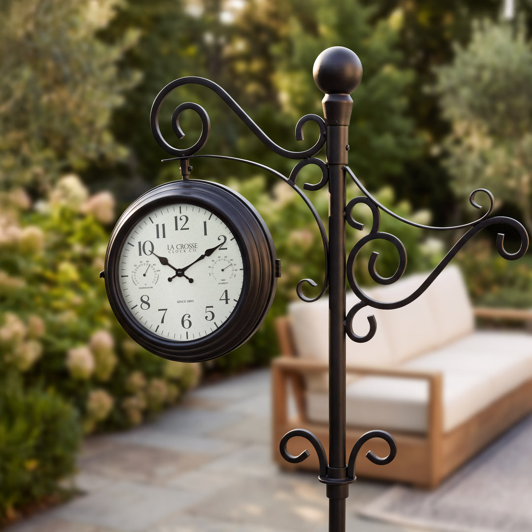 Lifestyle image of a close-up view of the 425-34188 Outdoor Dual-Sided Metal Pedestal Clock shown with a stone patio and outdoor funriture in the background. The clock features a temperature dial in a Fahrenheit scale, a humidity dial, decorative black metal hands, black numbers on a flecked white dial surrounded by a dark brown metal frame on a 72-inch decorative scrollwork pedestal.