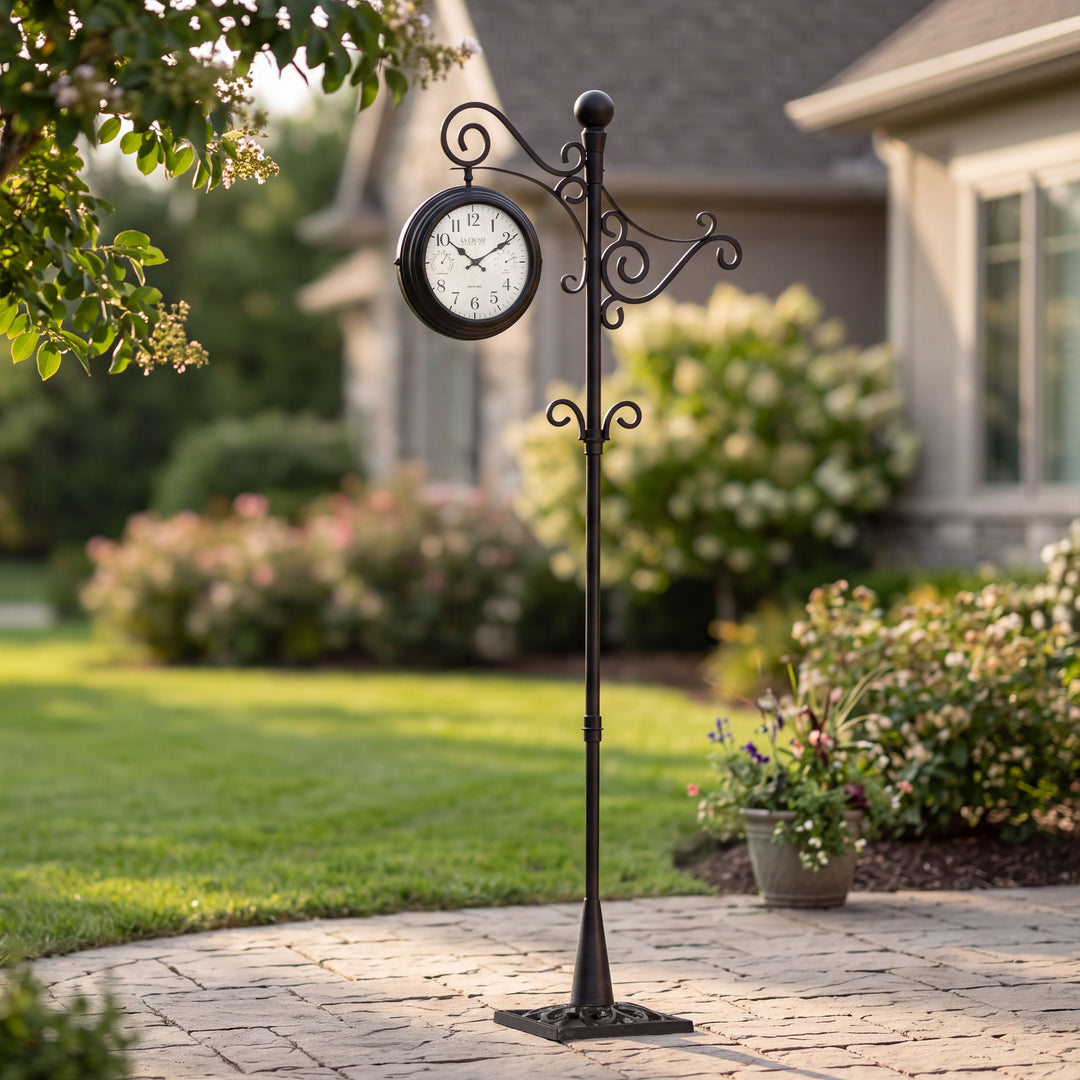 Lifestyle image of the 425-34188 Outdoor Dual-Sided Metal Pedestal Clock shown in front of a house with the yard and home in the background. The clock features a temperature dial in a Fahrenheit scale, a humidity dial, decorative black metal hands, black numbers on a flecked white dial surrounded by a dark brown metal frame on a 72-inch pedestal.