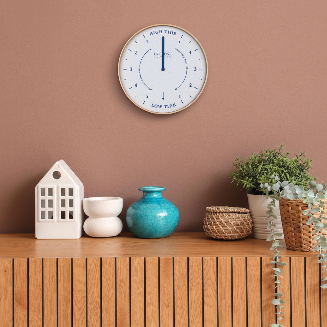 Lifestyle image of the 425-3831 11.8-inch Tide Wall Clock shown hanging on a terracotta wall above a wood slat side table with white and teal accessories. The clock features a light wood frame, a white and tan speckled dial, blue numbers, markings, and a blue metal hand.