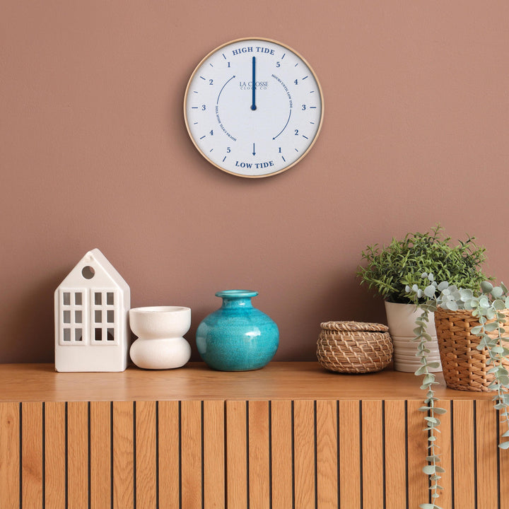 Lifestyle image of the 425-3831 11.8-inch Tide Wall Clock shown hanging on a terracotta wall above a wood slat side table with white and teal accessories. The clock features a light wood frame, a white and tan speckled dial, blue numbers, markings, and a blue metal hand.