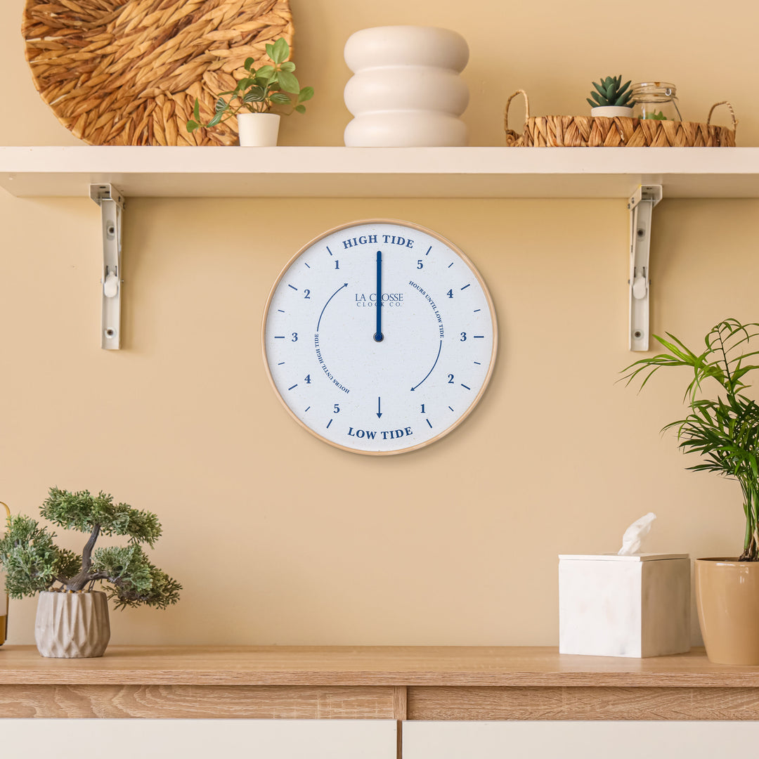 Lifestyle image of the 425-3831 11.8-inch Tide Wall Clock shown hanging on a tan wall above a wood side table with shelves and plants. The clock features a light wood frame, a white and tan speckled dial, blue numbers, markings, and a blue metal hand.