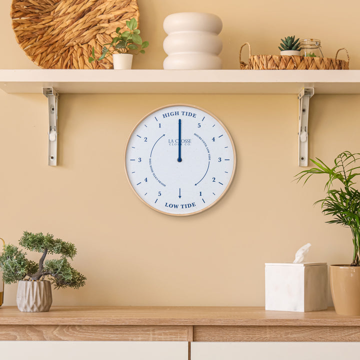 Lifestyle image of the 425-3831 11.8-inch Tide Wall Clock shown hanging on a tan wall above a wood side table with shelves and plants. The clock features a light wood frame, a white and tan speckled dial, blue numbers, markings, and a blue metal hand.