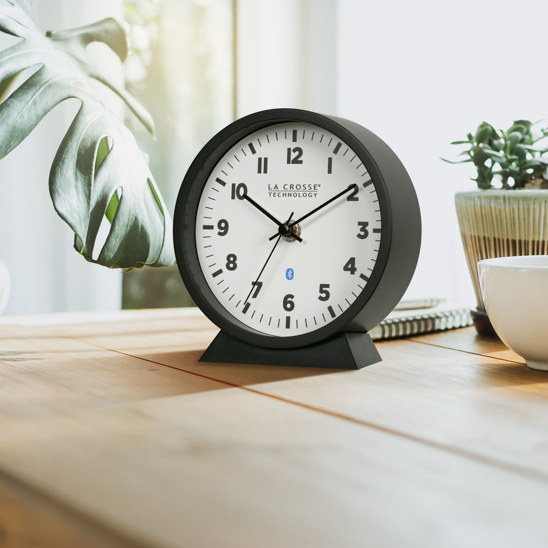 Lifestyle image of the 437-3816 Insta-Set Table/Wall Clock with Bluetooth® Technology shown sitting on a natural wood desk with plants and a coffee cup. The clock features a white dial with bold black numerals, black metal hands, and a black frame with a removable base for wall hanging.