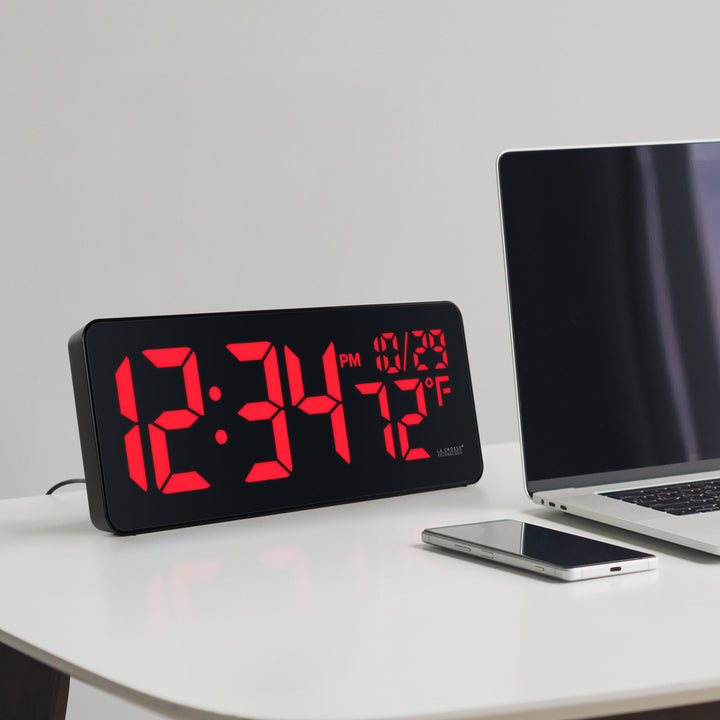 Lifestyle image of the 513-1411R Digital LED Wall Clock with Indoor Temperature shown sitting on a desk next to a laptop computer and a cell phone. The digital wall clock features large 3-inch LED time digits, indoor temperature, and the date.