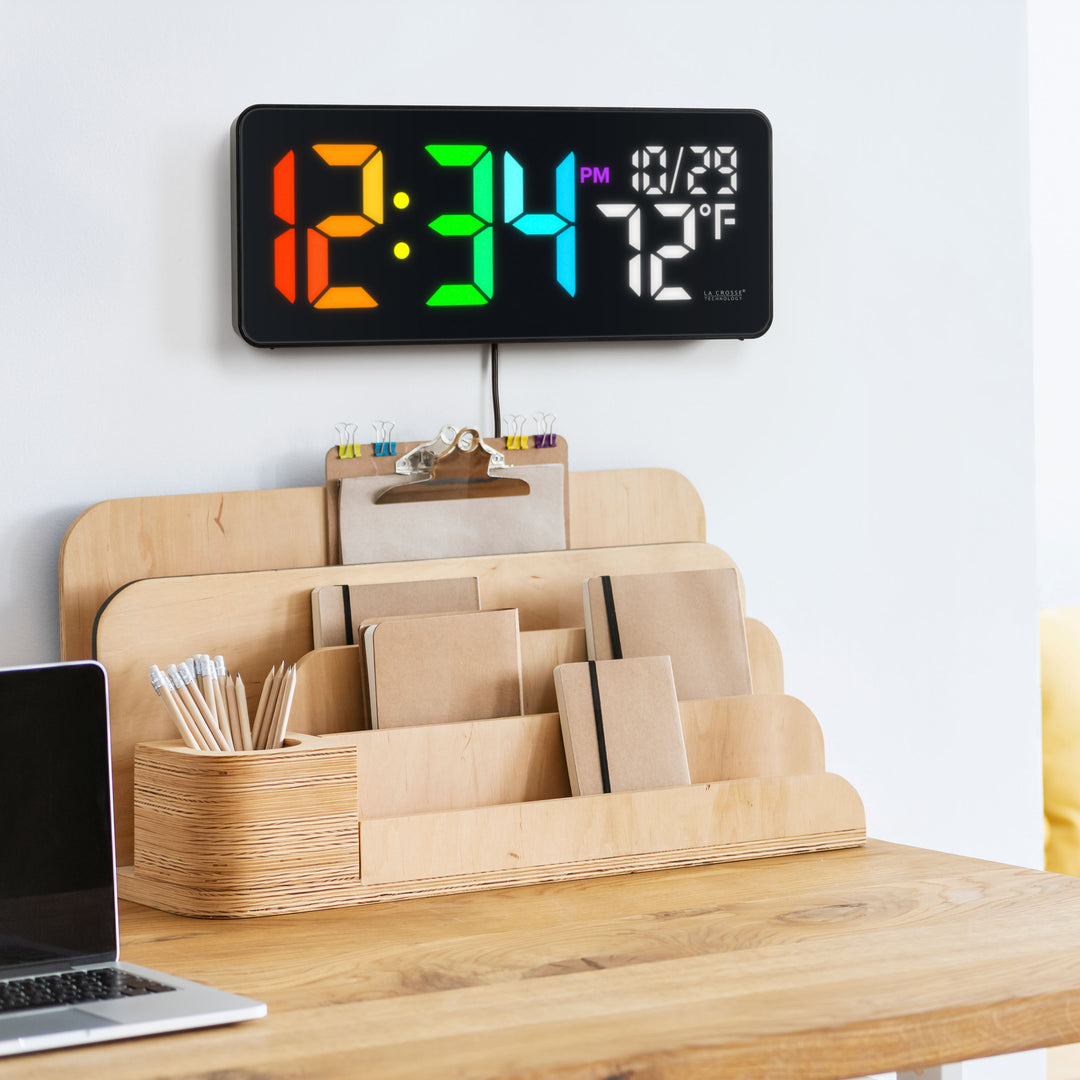 Lifestyle image of the 513-1411 Color-Changing LED Wall Clock hanging on a wall above a natural wood dek organizer in a sleek black case displaying time, date, and indoor temperature. Features 3-inch digits with a vibrant RGB-style color-changing display and a glossy lens finish.