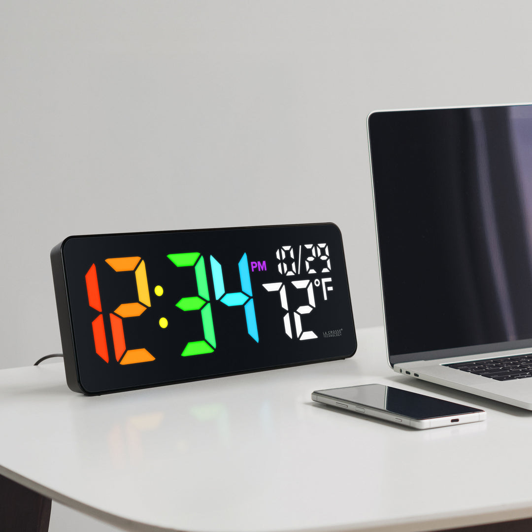 Lifestyle image of the 513-1411 Color-Changing LED Wall or Tabletop Clock sitting on a white desk next to a laptop computer and a phone with a black case displaying time in vibrant colors, date, and indoor temperature in white LED numbers. Features 3-inch digits with a vibrant RGB-style color-changing display and a glossy lens finish.