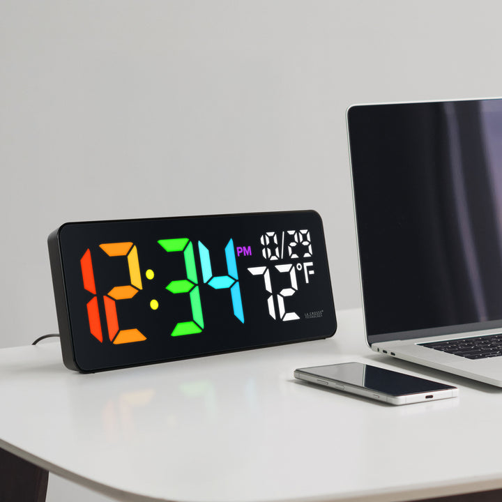 Lifestyle image of the 513-1411 Color-Changing LED Wall or Tabletop Clock sitting on a white desk next to a laptop computer and a phone with a black case displaying time in vibrant colors, date, and indoor temperature in white LED numbers. Features 3-inch digits with a vibrant RGB-style color-changing display and a glossy lens finish.