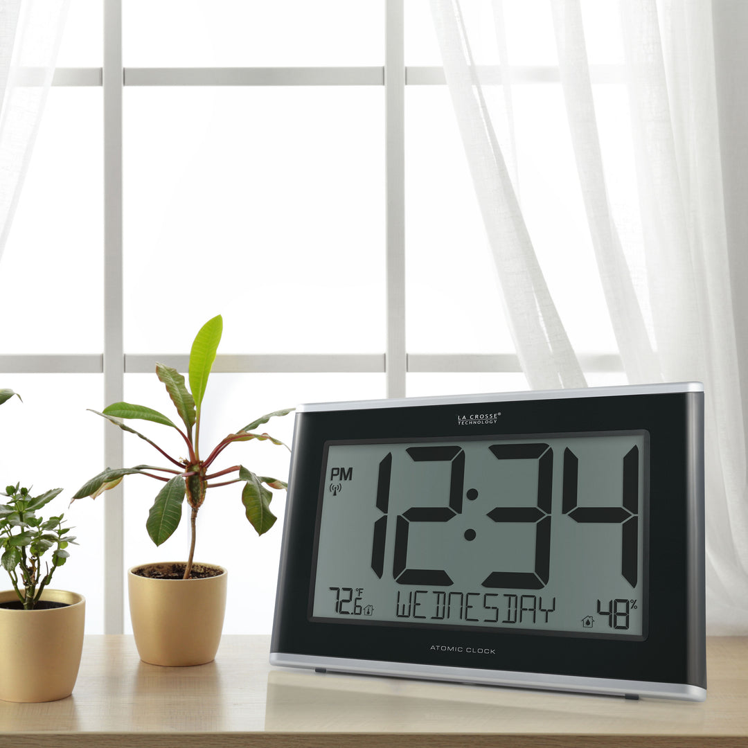 Lifestyle image of the 513-36908 Executive Atomic Digital Wall Clock with Temperature and Humidity sitting on a wood table in front of a window with wite sheers. The clock features a black frame, a large LCD screen displaying 12:34 pm, Wednesday, indoor temperature, humidity, and signal icons.