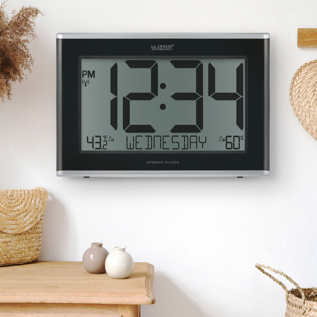 Lifestyle image of the 513-36908 Executive Atomic Digital Wall Clock with Temperature and Humidity hanging on a white wall above a light wood dresser. The clock features a black frame, a large LCD screen displaying 12:34 pm, Wednesday, indoor temperature, humidity, and signal icons.