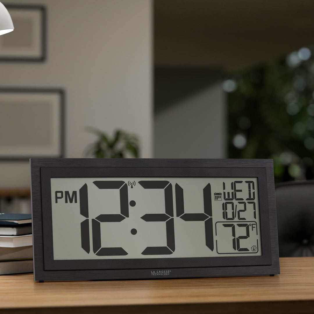Lifestyle image of the 515-1419v2 Atomic Digital Wall Clock with Time Advance shown on a wood desk in an office setting. The digital wall clock features a dual-textured black frame, easy-to-read 4.49-inch time digits, indoor temperature reading of 72 degrees Fahrenheit, and a calendar.