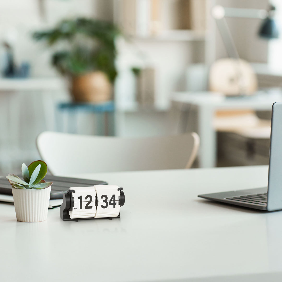 Lifestyle image of the 536-527W Rollins Mini Mechanical Flip Clock – White, shown sitting on a white desk next to a white potted plant with an office setting in the background. The clock features white hour and minute flip cards with black numbers in a black case.