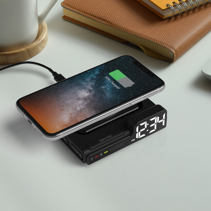 Lifestyle image of the 617-143 Amp Mini 15W Wireless Charging LED Alarm Clock shown on a desk from an aerial view with a phone sitting on the wireless charging pad. The alarm clock features easy access buttons, an alarm with snooze, white time digits, and LED status light shown here in red.