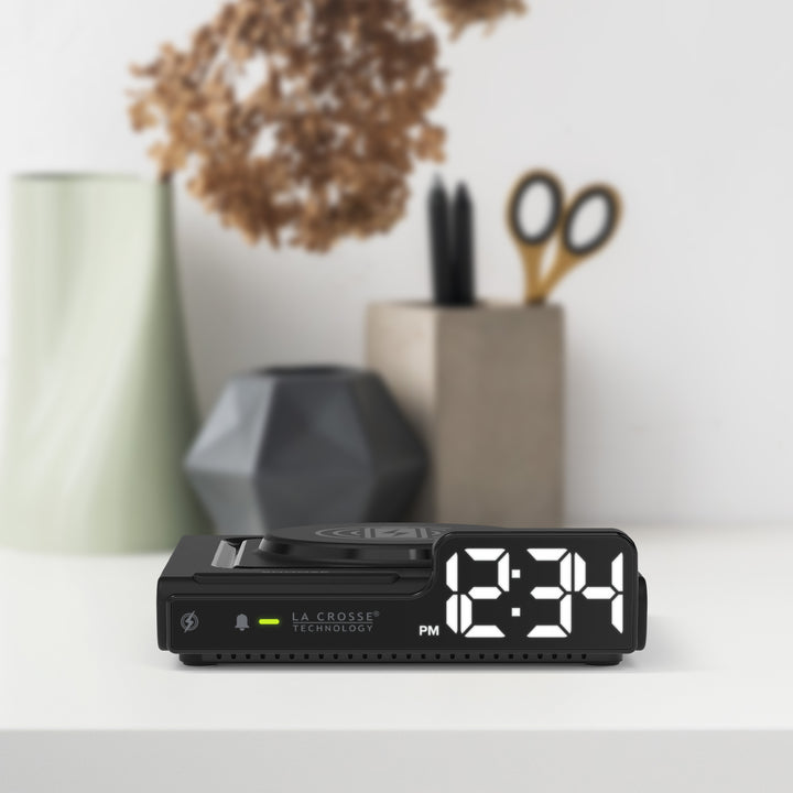Lifestyle image of the 617-143 Amp Mini 15W Wireless Charging LED Alarm Clock shown sitting on a white desk with desk accessories in the background. The alarm clock features a wireless charging pad, easy access buttons, an alarm with snooze, white time digits, and LED status light.