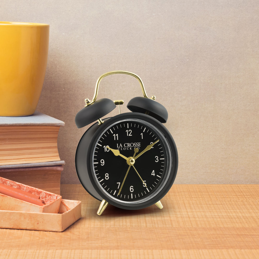 Lifestyle image of the 617-3314BG Metal Twin Bell Alarm Clock – Black, sitting on a light wood shelf with books. The clock features a black dial with white numbers, gold hands and accents, in a black metal case