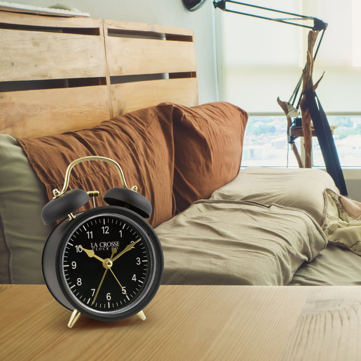 Lifestyle image of the 617-3314BG Metal Twin Bell Alarm Clock – Black, sitting on a nightstand with a bedroom scene in the background. The clock features a black dial with white numbers, gold hands and accents, in a black metal case.