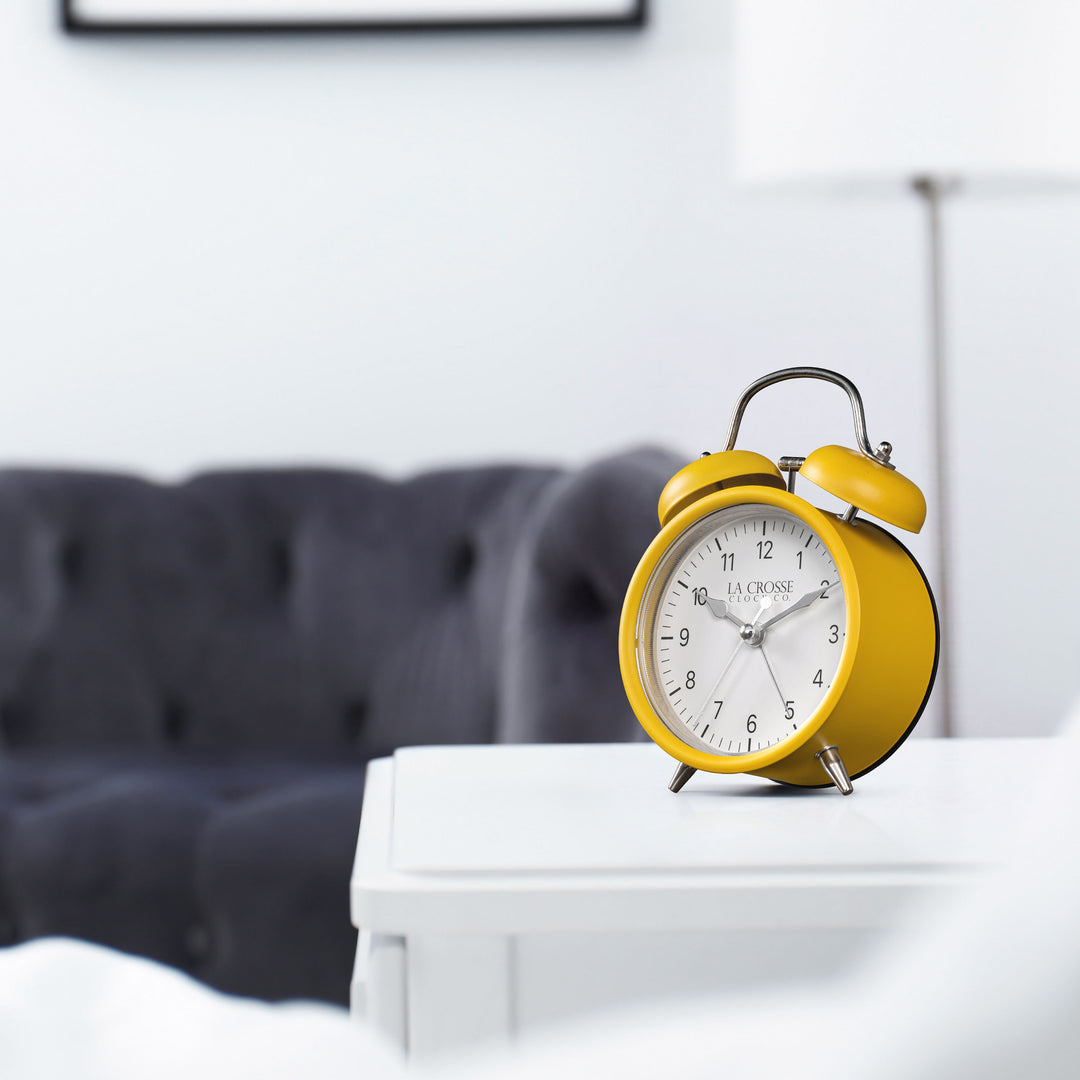 Lifestyle image of the 617-3314MY Metal Twin Bell Alarm Clock – Mustard Yellow, sitting on a white side table with a dark grey couch in the background. The clock features a white dial with black numbers, silver hands and accents, in a mustard yellow metal case.