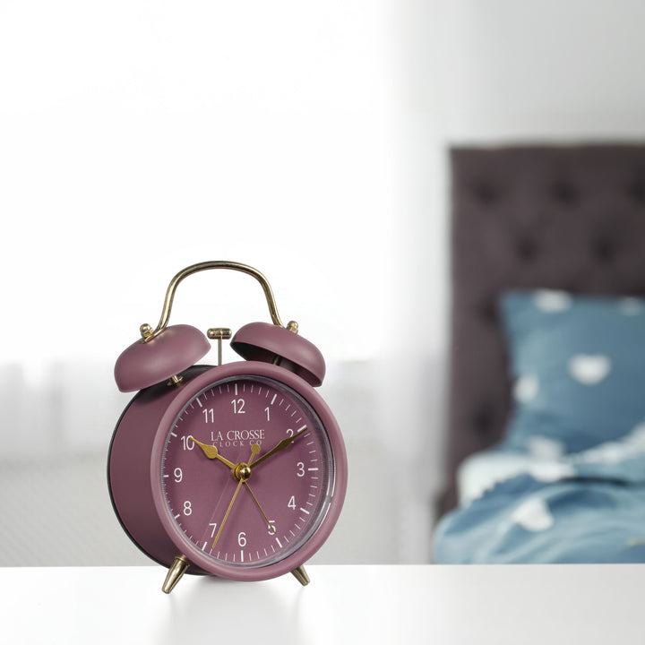 Lifestyle image of the 617-3314RR Metal Twin Bell Alarm Clock – Ruby Red, sitting on a nightstand with a bedroom scene in the background. The clock features a ruby red dial with white numbers, gold hands and accents, in a ruby red metal case.