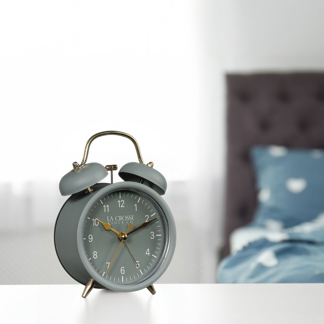 Lifestyle image of the 617-3314SG Metal Twin Bell Alarm Clock – Sage Green, sitting on a nightstand with a bedroom scene in the background. The clock features a sage green dial with white numbers, gold hands and accents, in a sage green metal case.