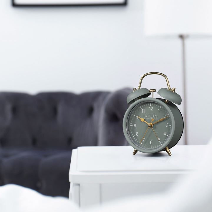 Lifestyle image of the 617-3314SG Metal Twin Bell Alarm Clock – Sage Green, sitting on a white side table with a dark grey couch in the background. The clock features a sage green dial with white numbers, gold hands and accents, in a sage green metal case.